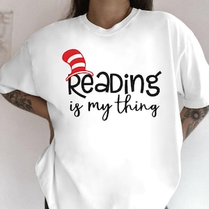 May include: White t-shirt with the text "Reading is my thing" in black script. The word "Reading" has a red and white striped hat above it. A casual, comfortable tee for book lovers.