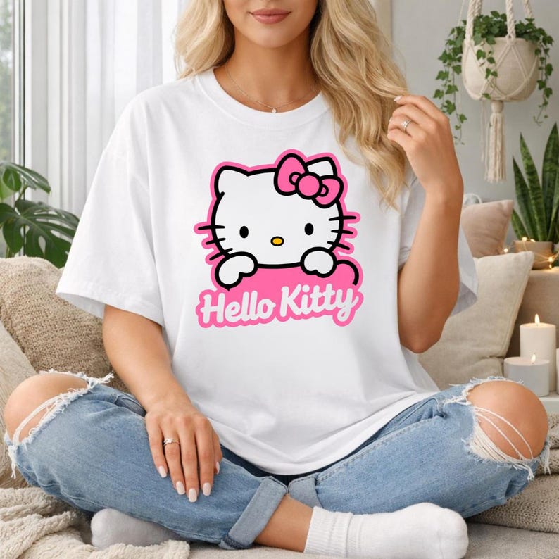 May include: White t-shirt featuring a Hello Kitty graphic. The graphic includes a white Hello Kitty face with a pink bow, whiskers, and the text "Hello Kitty" in pink. The shirt is worn with blue jeans.