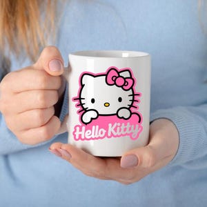 May include: White ceramic mug featuring a pink and black Hello Kitty design. The design includes Hello Kitty's face, a pink bow, and the words "Hello Kitty" in pink. The mug is held by a person wearing a blue long-sleeve shirt.