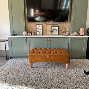May include: A brown tufted storage ottoman with wooden legs sits in front of a green cabinet with a white countertop. The cabinet has a television mounted above it and a decorative wooden wall behind it.