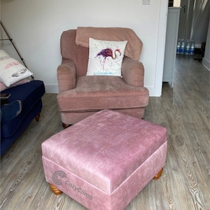 May include: A pink upholstered armchair with a throw blanket and a cushion featuring a watercolour flamingo design. A matching pink footstool with wooden legs sits in front of the chair.