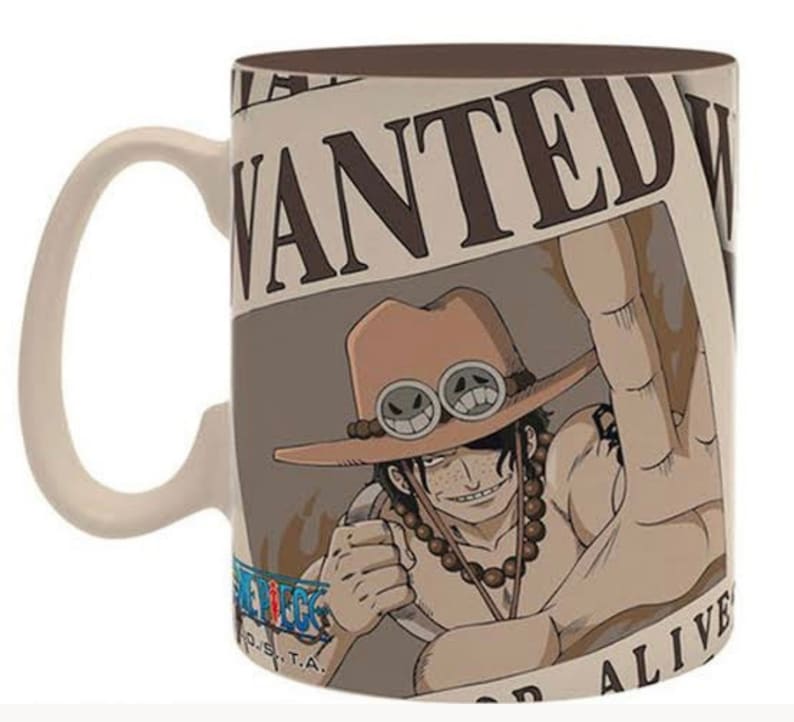 May include: A white ceramic mug with a brown handle. The mug features a wanted poster with a cartoon image of a man with brown hair, a brown hat with goggles, and a beaded necklace. The poster says "WANTED" at the top and "DEAD OR ALIVE" at the bottom.