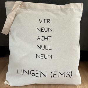 Könnte beinhalten: Cremefarbene Tragetasche mit schwarzem Text. Der Text lautet "VIER NEUN ACHT NULL NEUN LINGEN (EMS)". Die Tasche hat ein schlichtes Design mit rechteckiger Form und Griff aus gleichem Material.