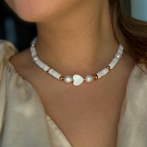 Könnte beinhalten: Eine Halskette aus weißen Muscheln und goldenen Perlen mit einem weißen Herz-Anhänger.