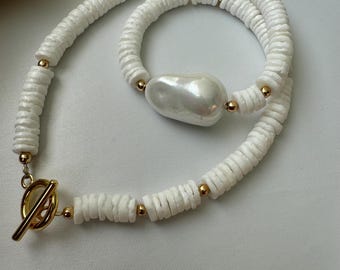 Op de afbeelding: Een witte schelpketting met een grote witte parel hanger en gouden accenten. De ketting is gemaakt van kleine, platte, witte schelpen die aan elkaar geregen zijn op een gouden ketting.