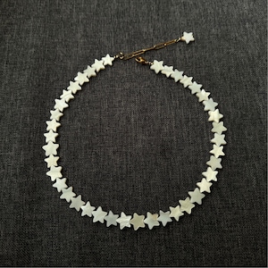 May include: A delicate necklace with small, star-shaped beads. The beads are pearly white and form a choker. The clasp and chain are gold-coloured. The necklace is displayed on a dark grey background.