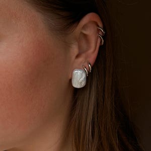 Puede incluir: Primer plano de una oreja adornada con un gran pendiente de perla blanca cuadrada. La oreja también presenta tres pendientes de aro plateados. El pendiente de perla tiene un aspecto veteado. El fondo es borroso y de tono neutro.