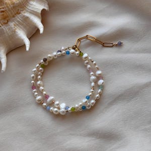 May include: A double-strand pearl bracelet with colorful beads and a gold chain clasp. The bracelet features white pearls interspersed with pink, blue, green, and gold beads. The clasp is a gold chain with a small blue bead at the end.