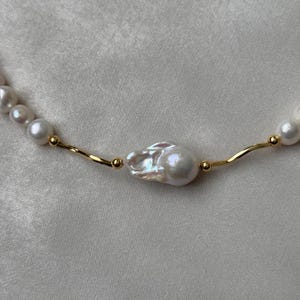 Peut inclure: Gros plan sur un collier de perles avec une chaîne en or. Le collier présente une grosse perle de forme irrégulière comme point central, avec de plus petites perles rondes et des détails dorés. Le fond est un tissu blanc cassé.