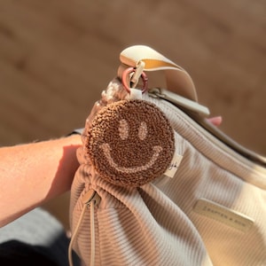 Könnte beinhalten: Ein runder, brauner, getufteter Smiley-Schlüsselanhänger mit cremefarbenem, gesticktem Lächeln und Augen, befestigt an einer beigen Cordtasche mit einem kupferfarbenen Metallring. Ein kleines weißes Stoffetikett mit schwarzem Text ist seitlich sichtbar.