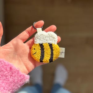 Könnte beinhalten: Ein Punch-Needle-Schlüsselanhänger in Form einer Biene mit gelb-schwarz gestreiftem Körper, weißen Flügeln und einem kleinen gestickten Lächeln. Der Schlüsselanhänger hat einen silbernen Metallring und ein kleines Stoffetikett mit schwarzem Text.