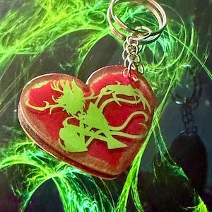 May include: Heart-shaped keychain with a red background and a green silhouette design. The keychain has a silver-colored metal ring and chain. The design appears to be a stylized image of a person or creature.