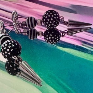May include: A pair of earrings with black and white striped and dotted beads, linked by silver-toned metal chains. Each earring has a conical, silver-toned spike at the bottom. The earrings are displayed on a reflective, iridescent surface.