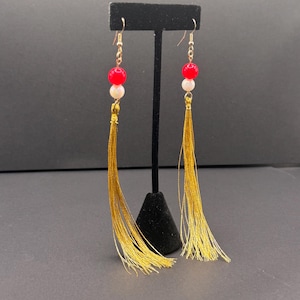May include: A pair of dangling earrings with red and white beads and long gold tassels. The earrings are displayed on a black stand against a dark background. The earrings have gold-colored hooks.
