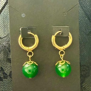 Könnte beinhalten: Ein Paar goldfarbene Creolen mit einem hängenden grünen Orb. Der Orb hat eine leuchtend grüne Farbe und eine dekorative goldfarbene Kappe. Die Ohrringe werden auf einer schwarzen Karte präsentiert.