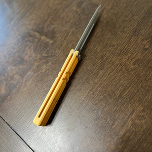 May include: A yellow and silver folding knife with a blade that is closed. The knife is on a wooden surface.