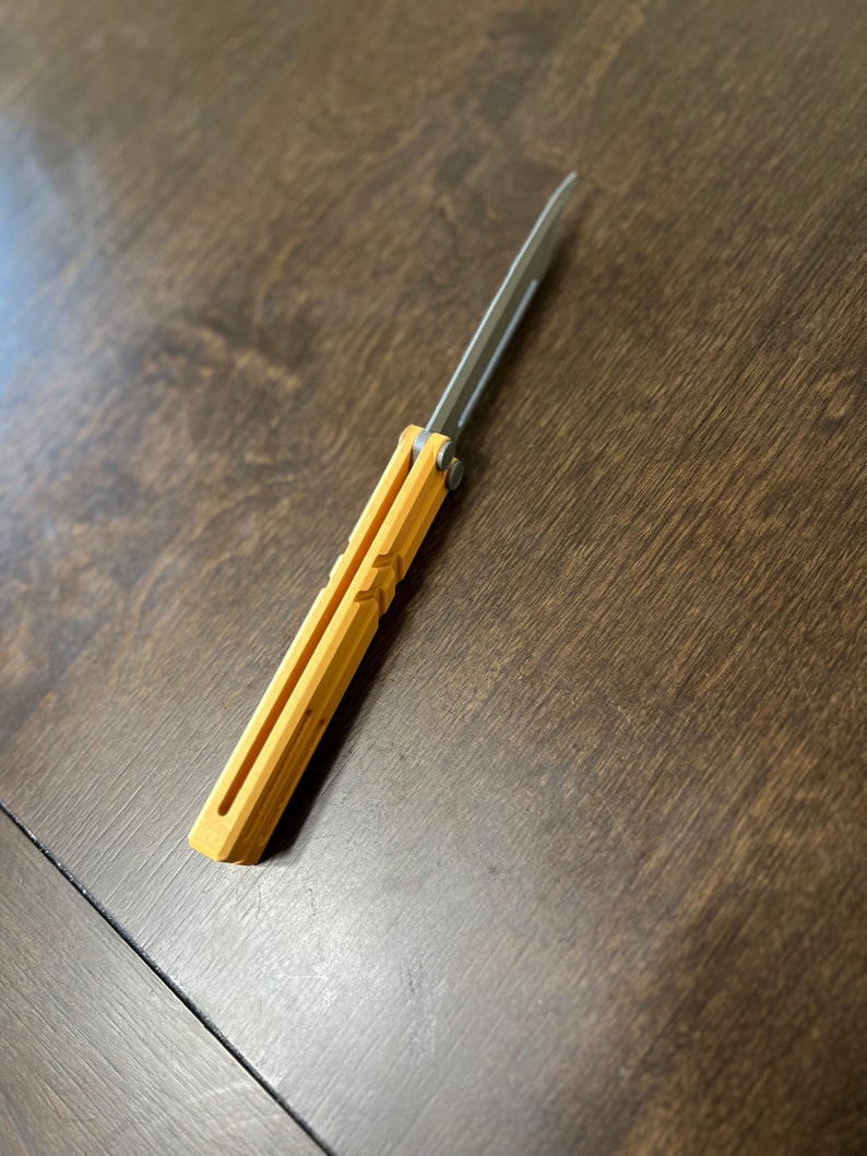 May include: A yellow and silver folding knife with a blade that is closed. The knife is on a wooden surface.