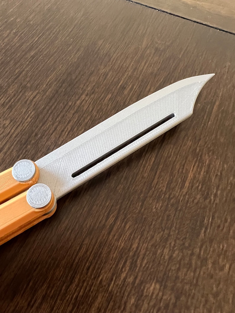May include: A silver butterfly knife with a textured blade and orange handles. The knife is partially open, showing the mechanism.