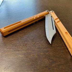 May include: A brown and silver butterfly knife with a textured blade. The knife is open and the blade is partially extended.