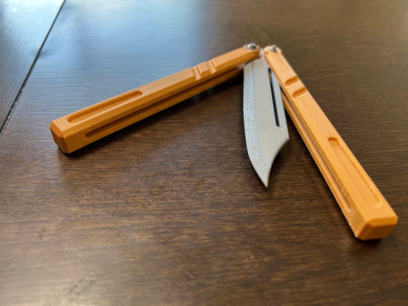 May include: A brown and silver butterfly knife with a textured blade. The knife is open and the blade is partially extended.