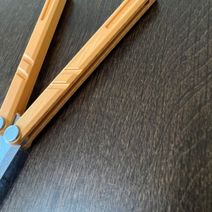 May include: A brown wooden butterfly knife with a silver metal hinge. The knife is closed and lying on a dark brown wooden surface.
