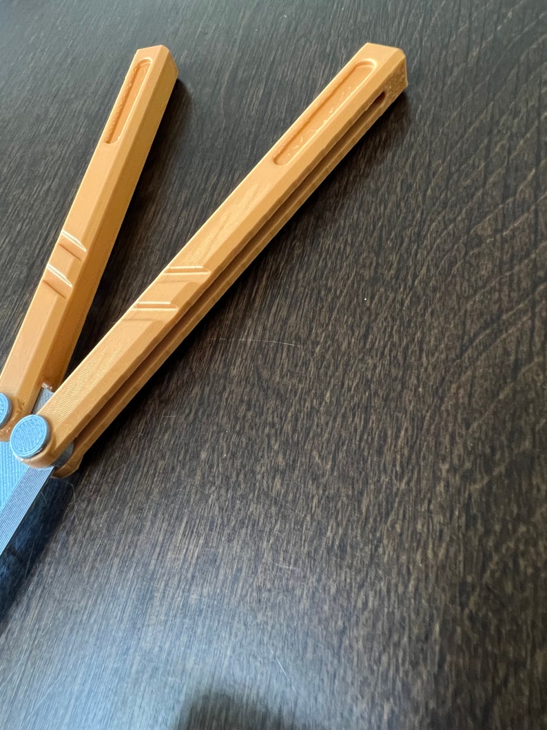 May include: A brown wooden butterfly knife with a silver metal hinge. The knife is closed and lying on a dark brown wooden surface.