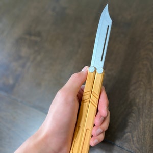 May include: A wooden butterfly knife with a silver blade. The knife is closed and being held in a hand.