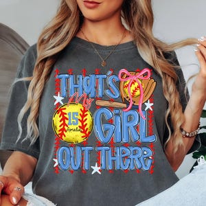 May include: Dark gray t-shirt with a softball-themed graphic. The design includes a softball, glove, and the text "That's My Girl Out There." The softball has the number 15 and the name "Amelia" on it. The graphic is in blue, red, yellow, and pink.