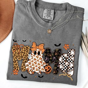 May include: A charcoal gray t-shirt featuring a graphic that spells MAMA, where the first letter is leopard print, the second is a ghost with a bow, the third has a pumpkin pattern, and the fourth is a black and white checkerboard with ghosts.