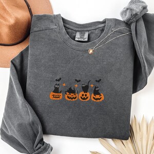 May include: Dark gray sweatshirt with an embroidered Halloween design. The design features four orange pumpkins with black cats and bats. The sweatshirt has long sleeves and a crew neck. A gold necklace is visible.