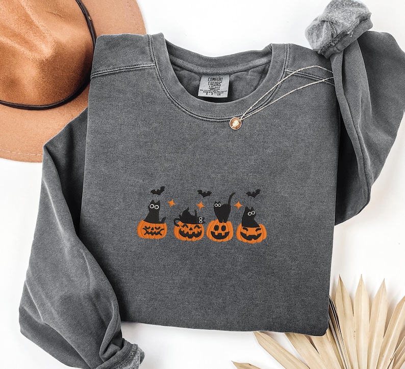 May include: Dark gray sweatshirt with an embroidered Halloween design. The design features four orange pumpkins with black cats and bats. The sweatshirt has long sleeves and a crew neck. A gold necklace is visible.