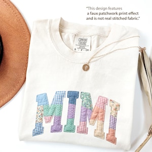 May include: Cream-colored t-shirt with the word "MIMI" in patchwork-style letters. The letters are in various pastel colors and patterns. A gold necklace with a pendant is draped on the shirt. The shirt has a tag that says "COMFORT COLORS".