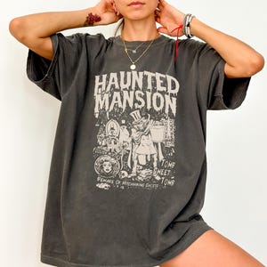 May include: A charcoal gray short-sleeve t-shirt featuring a cream-colored graphic print with the text Haunted Mansion, Foolish Mortals, Tomb Sweet Tomb, and Beware of Hitchhiking Ghosts, depicting various characters and a top-hatted figure.