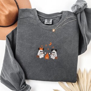 May include: A charcoal gray crewneck sweatshirt featuring an embroidered graphic of two ghosts with mouse ears roasting marshmallows over a campfire. The design includes orange stars and a crescent moon. The inner neck label reads Comfort Colors.