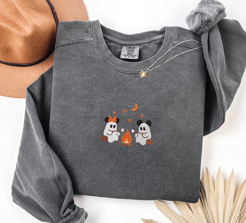 May include: A charcoal gray crewneck sweatshirt featuring an embroidered graphic of two ghosts with mouse ears roasting marshmallows over a campfire. The design includes orange stars and a crescent moon. The inner neck label reads Comfort Colors.