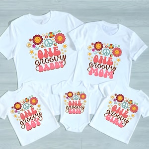 May include: Set of five white t-shirts with a groovy 70s theme. The shirts feature a peace sign, flowers, and the text "One groovy [name]" in pink and brown lettering.