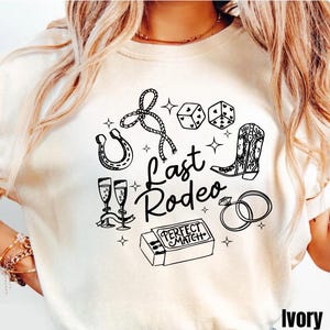 May include: Ivory-colored t-shirt with a black graphic design featuring a horseshoe, dice, cowboy boot, champagne glasses, and the words "Last Rodeo." The phrase "Perfect Match" is on a matchbox, and there are two rings.