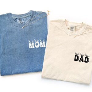 May include: Two t-shirts, one blue and one cream-colored, are displayed. The blue shirt has the word "MOM" printed in white with bunny ears, and the cream shirt has "DAD" printed in black with bunny ears. Both shirts have a necklace.