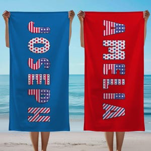 May include: Two beach towels, one blue and one red, are held up against a beach and ocean backdrop. The blue towel says "JOSEPH" in large, patriotic letters. The red towel says "AMELIA" in similar letters, both with a stars and stripes design.