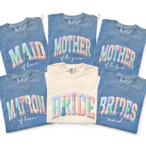 May include: A collection of blue and cream-colored t-shirts with text for a wedding party. The shirts feature the words "MAID of honor", "MOTHER of the groom", "MOTHER of the bride", "BRIDE", "MATRON of honor", and "BRIDES maid" in a patchwork design.