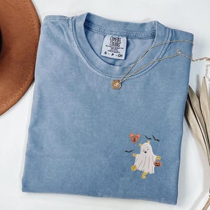 May include: A blue Comfort Colors t-shirt with a round neckline. The shirt features an embroidered design of a ghost dressed as Winnie the Pooh, holding a pumpkin. A gold necklace with a pendant is draped across the shirt.