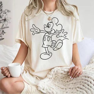 May include: A white t-shirt with a black outline of Mickey Mouse in a walking pose. The shirt is worn by a person sitting on a white blanket.