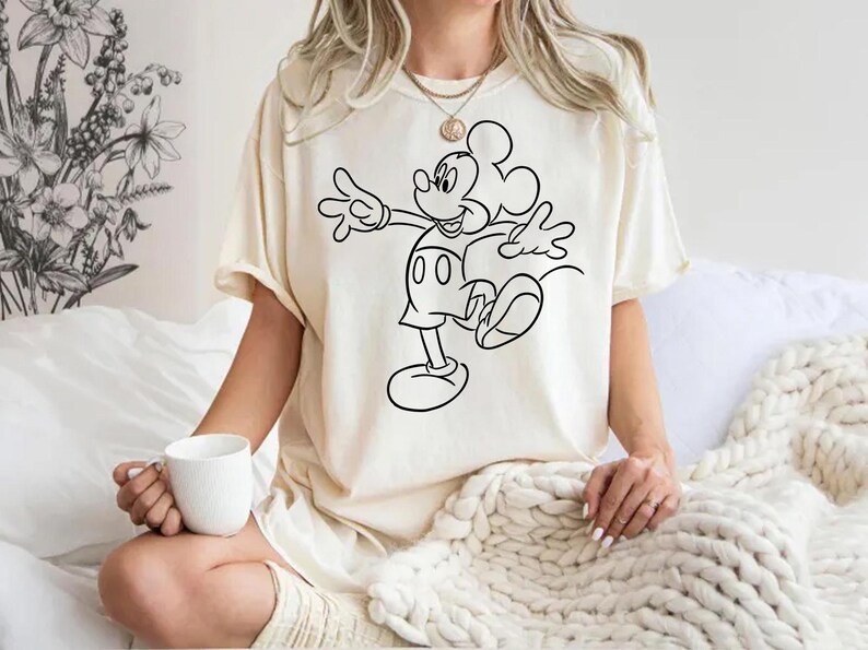 May include: A white t-shirt with a black outline of Mickey Mouse in a walking pose. The shirt is worn by a person sitting on a white blanket.