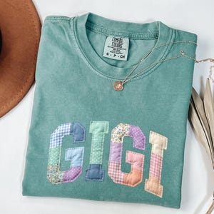 May include: A teal Comfort Colors t-shirt with the name "GIGI" appliqued in patchwork fabric. The letters are in various colors and patterns. A silver necklace with a small pendant rests on the shirt.