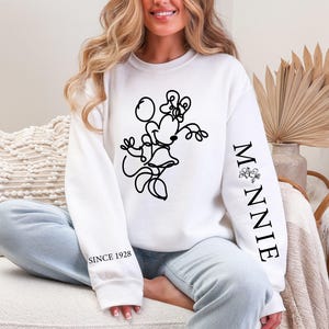 May include: White sweatshirt featuring a black line drawing of Minnie Mouse. The sleeve reads "MINNIE" with a floral accent, and "SINCE 1928" is printed on the other sleeve. The sweatshirt is a casual style.