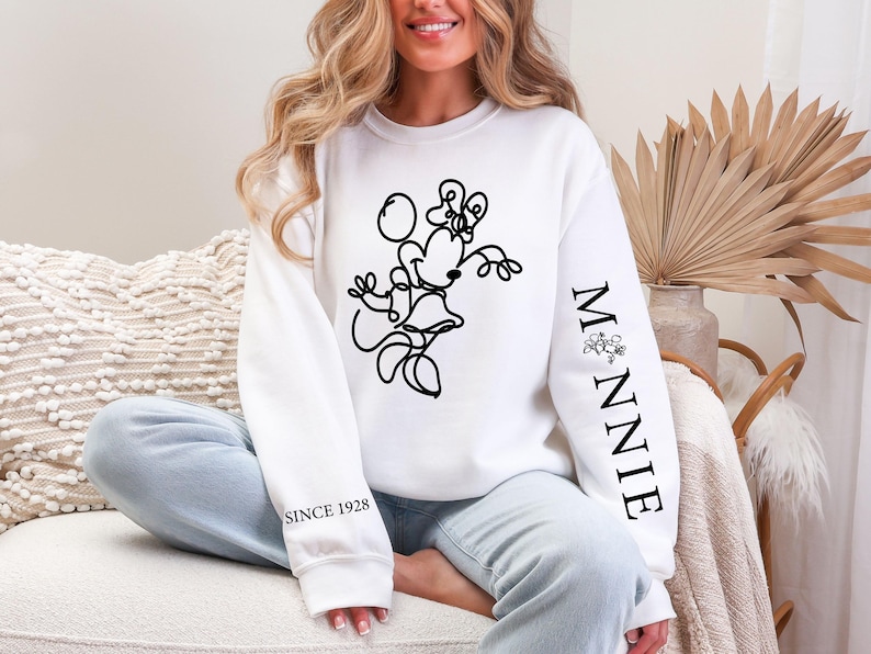 May include: White sweatshirt featuring a black line drawing of Minnie Mouse. The sleeve reads "MINNIE" with a floral accent, and "SINCE 1928" is printed on the other sleeve. The sweatshirt is a casual style.