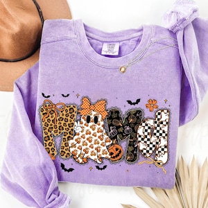 May include: A light purple crewneck sweatshirt featuring a graphic that spells MAMA. The letters are decorated with leopard print, a ghost wearing a bow, pumpkin patterns, and a black and white checkerboard pattern with small ghost silhouettes.