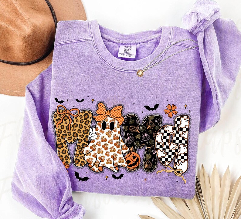 May include: A light purple crewneck sweatshirt featuring a graphic that spells MAMA. The letters are decorated with leopard print, a ghost wearing a bow, pumpkin patterns, and a black and white checkerboard pattern with small ghost silhouettes.