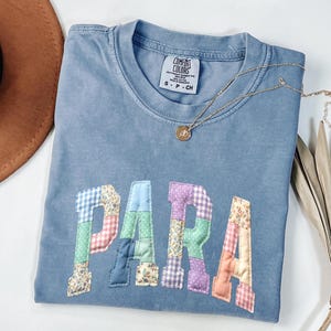 May include: A blue t-shirt with the word "PARA" appliquéd in patchwork fabric. The letters are in various floral and patterned fabrics. A gold necklace with a pendant is on the shirt. The shirt is labeled "Comfort Colors".