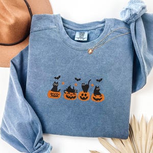 May include: A blue crewneck sweatshirt featuring an embroidered Halloween design. The design includes four black cats sitting on orange pumpkins with bats flying above. A gold necklace is draped on the sweatshirt.